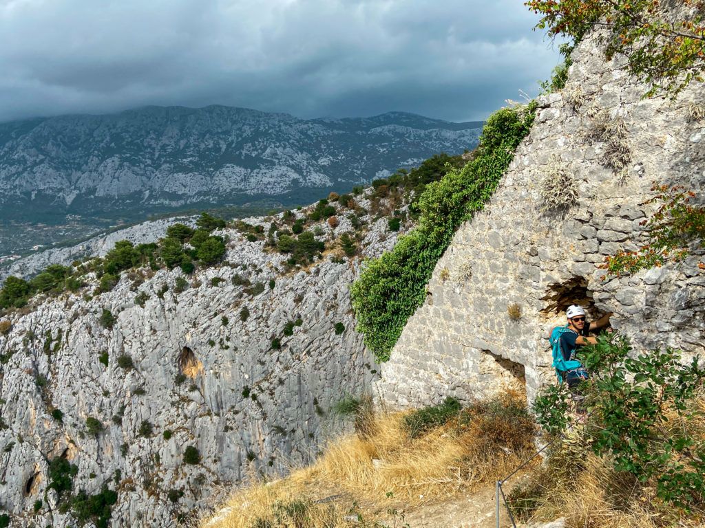 Marcin przechodzi przez dziurę w murze na samym końcu ferraty Fortica w Omiš , za nim w tle góry