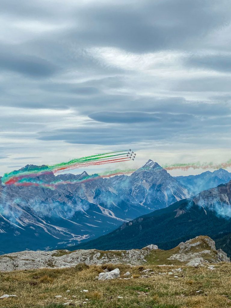 Pokaz sił powietrznych Italii podczas manewrów w okolicach Przełęczy Falzarego. Samoloty odrzutowe lecące w szyku, a za nimi ciągnąca się wstęga w kolorach falgi włoskiej. 