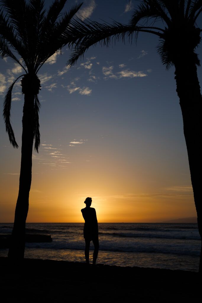 Ewa stoi między palmami podczas zachodu słońca na Playa de las Americas na Teneryfie